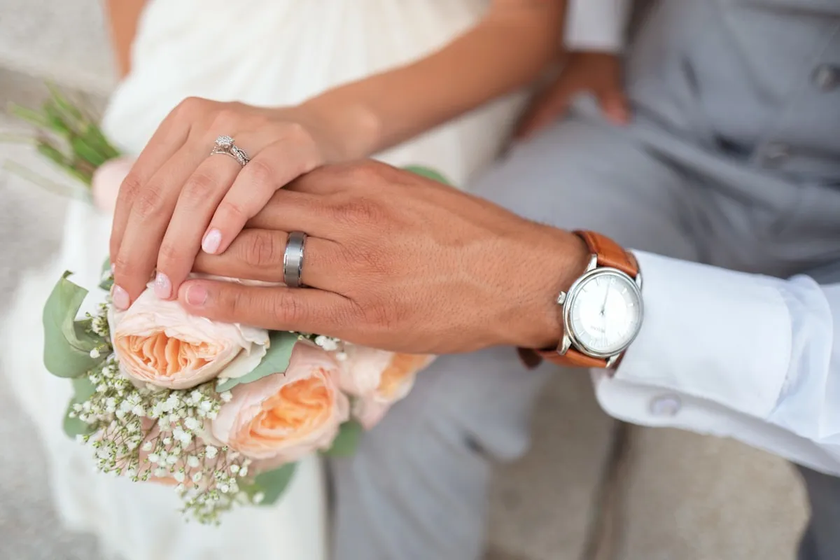 Mãos dos noivos com as alianças, apoiadas sobre um buquê de rosas